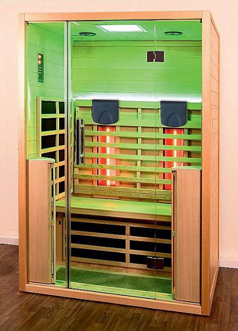 Die Infrarotsauna "Sonder-Edition" mit einer Glasfront und Rahmen aus hellem Hemlock-Holz. Im Inneren dieser 2-Personen Gesundheits- und Entspannungskabine befinden sich hinter einem ergonomisch geformten Anlehngitter zwei rötlich leuchtende Rückenstrahler, darüber im oberen Teil des Anlehngitters zwei schwarze Kopfstützen. Die Kabine steht auf einem dunklen Holzboden vor einer hellen Wand.
